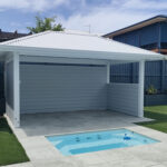 White Pool Cabana with white VJ ceiling, grey Hardie Linea cladded walls and a half wall it is set behind the in ground spa and looks out over the pool area.