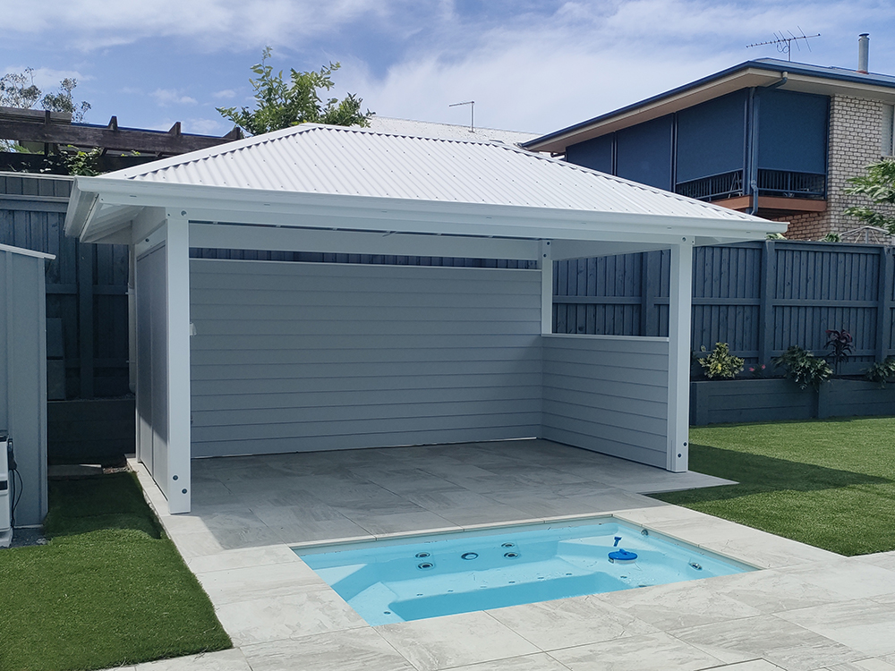 White Pool Cabana with white VJ ceiling, grey Hardie Linea cladded walls and a half wall it is set behind the in ground spa and looks out over the pool area.