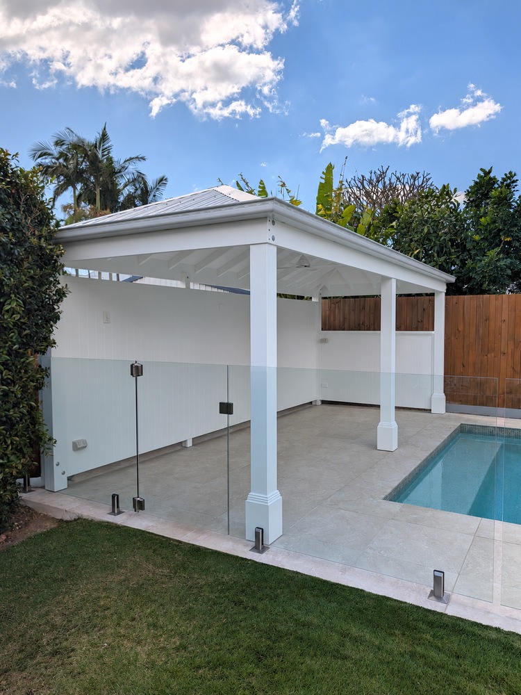 Corner view of the white hip roof Pool Cabana beside the tiled pool surround and glass fence.