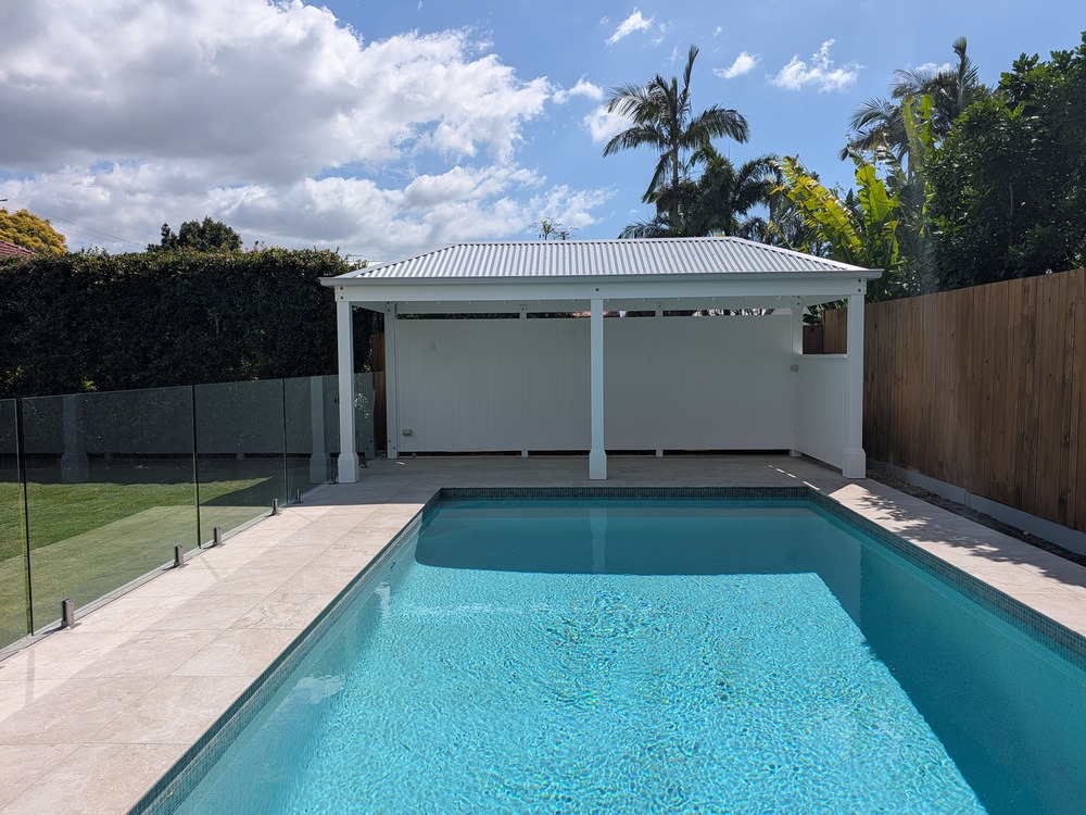 White hip roof Pool Cabana viewed across the swimming pool, with a full rear wall behind the front posts.