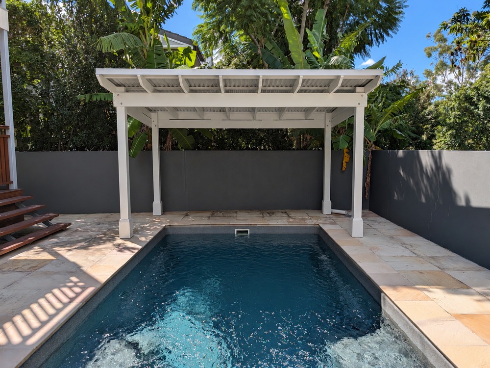 Front view across the swimming pool toward a white Roofed Pergola with a skillion roof and four timber posts.